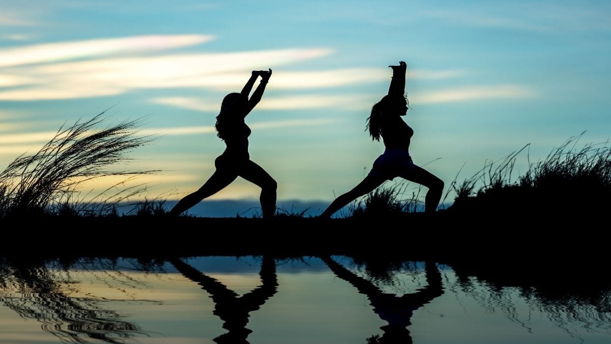 "Two silhouetted figures practicing hatha yoga: 21 powerful poses at sunset, standing on grass with their reflections visible in the calm water below, under a sky with soft clouds."