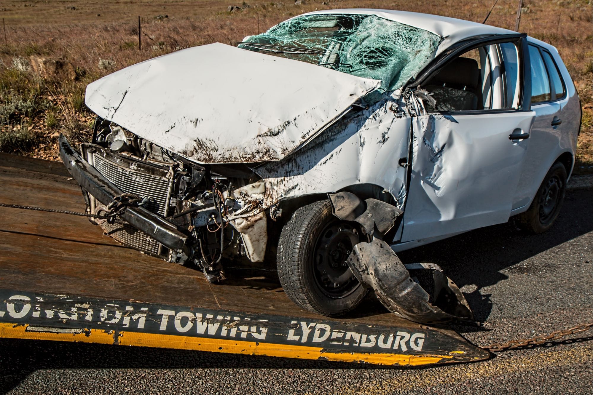 "A severely damaged white car with a shattered windshield and crumpled front end being towed on a flatbed truck labeled 'Lydendburg Towing,' set against a rural roadside background with dry grass and a clear sky."