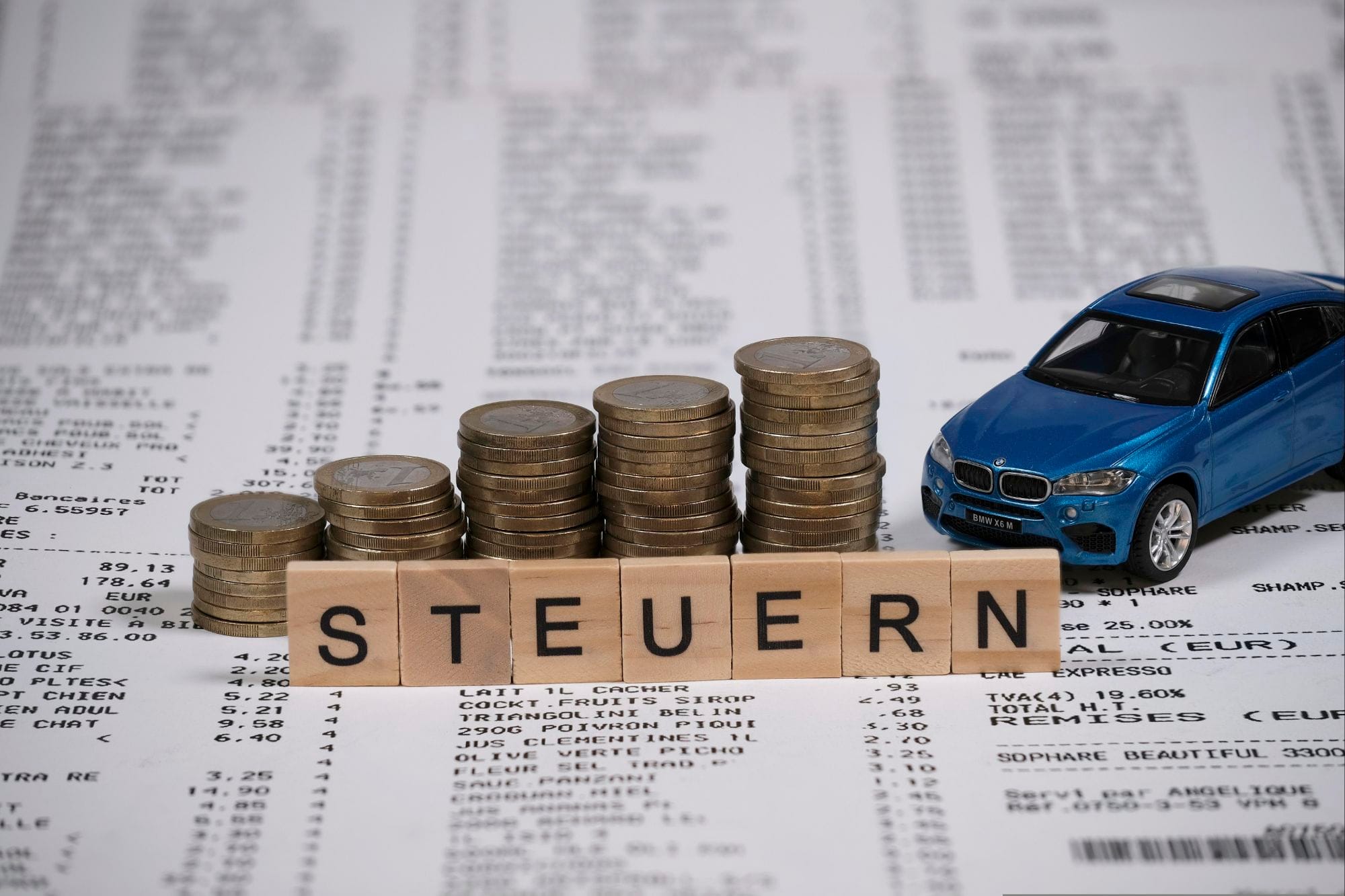 "A photograph featuring a blue toy car placed on a financial document with stacks of coins in the foreground. Wooden letter blocks spell out 'STEUERN' (likely intended as 'Steuer,' the German word for tax). The document appears to be a receipt or invoice with various itemized amounts and currency in euros, suggesting a focus on car-related expenses or taxes."