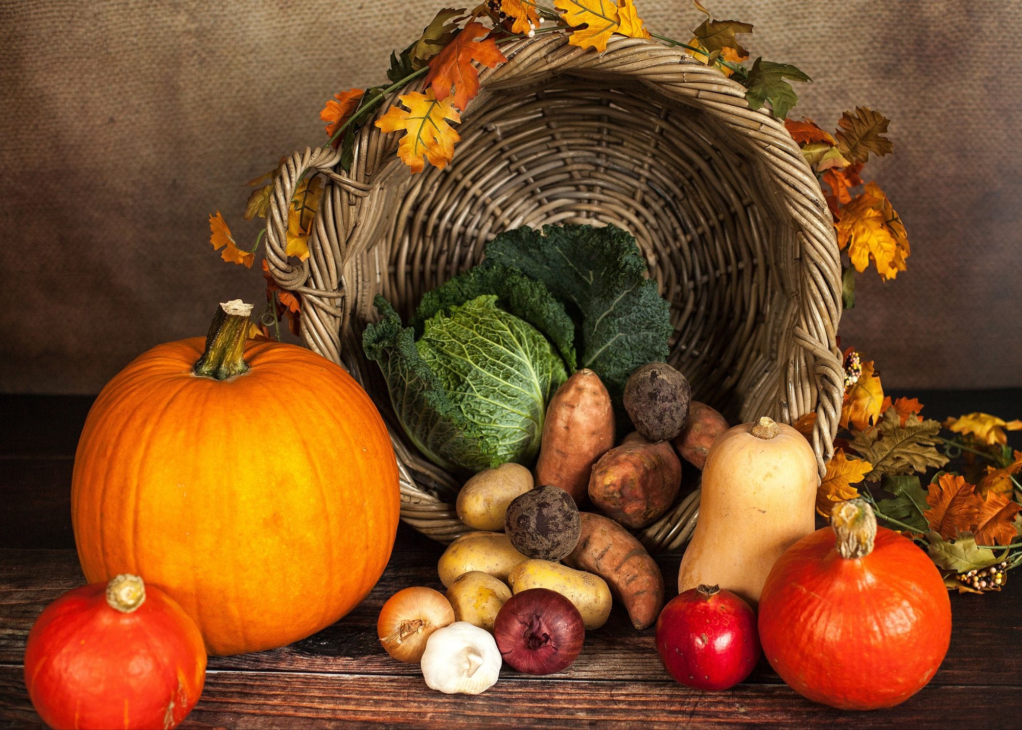 Fibermaxxing: 10 Powerful Steps to Transform You.
A rustic wooden table displays a harvest-themed still life with a woven basket filled with autumn leaves and various vegetables. The basket contains cabbage, potatoes, sweet potatoes, onions, garlic, and a butternut squash. Surrounding the basket are large pumpkins, a small red fruit, and additional autumn foliage, creating a warm, seasonal scene.