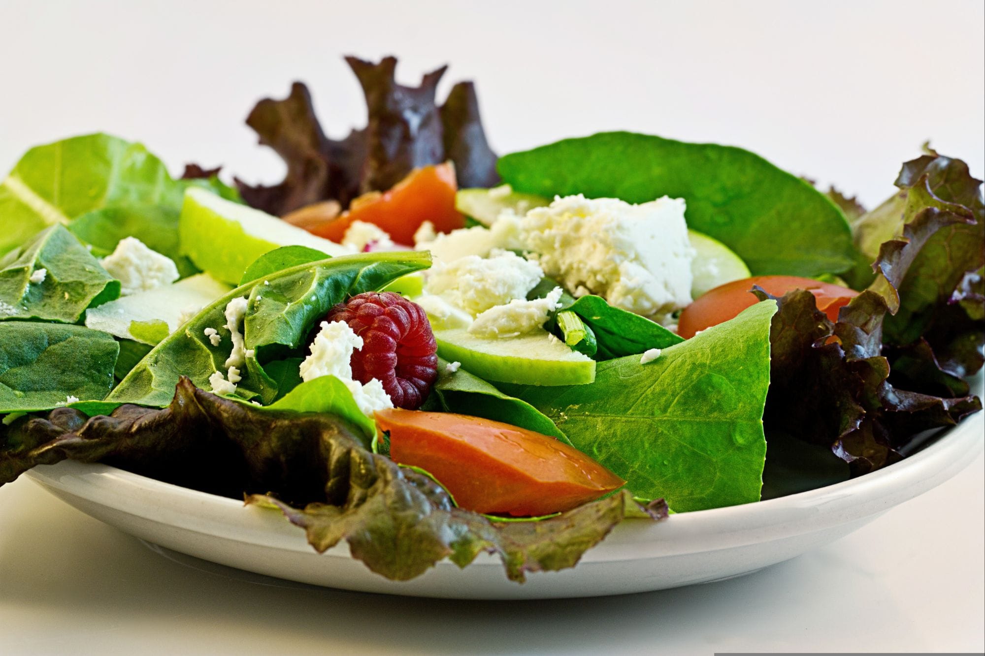 Fibermaxxing: 10 Powerful Steps to Transform You.
A white plate holds a fresh salad with green leafy lettuce, spinach, sliced tomatoes, cucumber, apple, raspberries, and crumbled white cheese, presented against a plain background.
