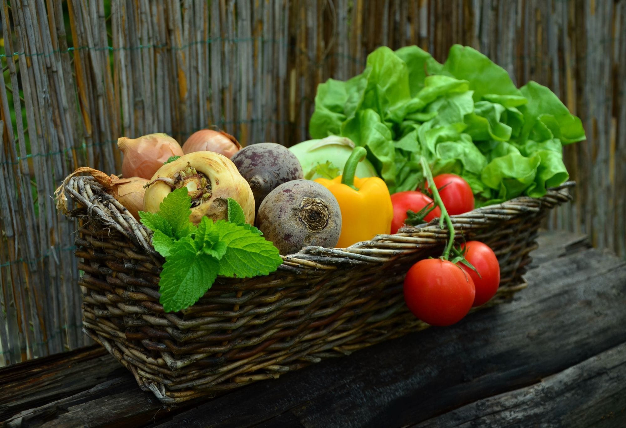 Fibermaxxing: 10 Powerful Steps to Transform You.
A woven basket on a wooden surface contains a variety of fresh vegetables and herbs, including lettuce, tomatoes, onions, beets, a yellow bell pepper, turnips, and mint leaves, set against a rustic bamboo fence background.