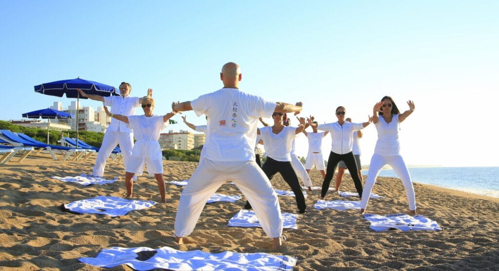 Hatha yoga: 21 powerful 
"A group of people in white clothing practicing a synchronized exercise or yoga pose on a sandy beach, with blue umbrellas and lounge chairs in the background and the sea visible on the horizon under a clear sky."